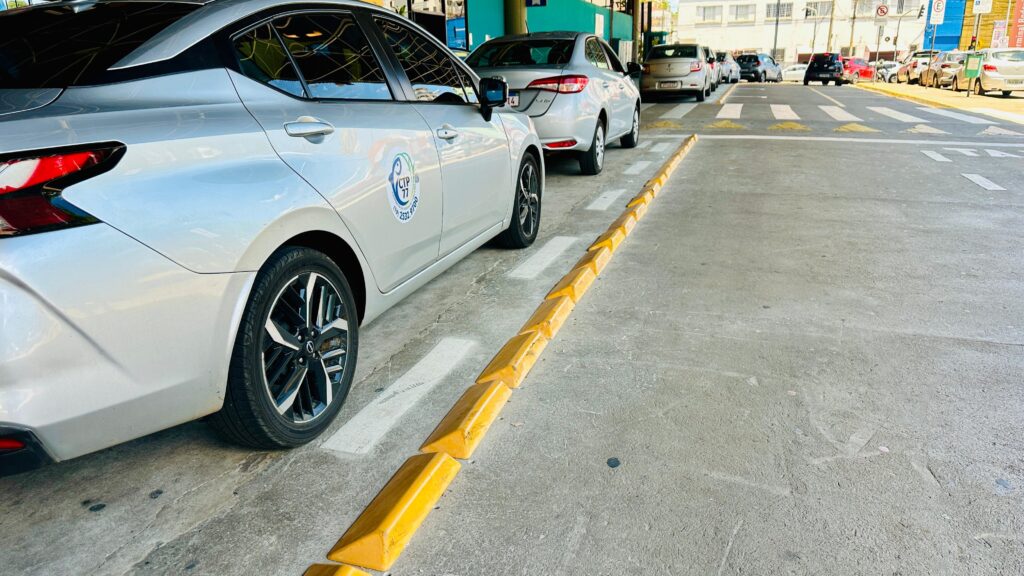 A imagem mostra uma fila de carros estacionados em um ambiente urbano, possivelmente um estacionamento ou uma rua. No primeiro plano, vemos a lateral de um carro prateado, com detalhes da roda e da traseira. Uma linha de tachões amarelos delimita a área de estacionamento, separando-a da via. Mais adiante, outros carros prateados e de outras cores seguem na fila. Ao fundo, é possível observar a arquitetura de edifícios e sinais de trânsito, indicando que se trata de uma área movimentada. A iluminação sugere que a foto foi tirada durante o dia.