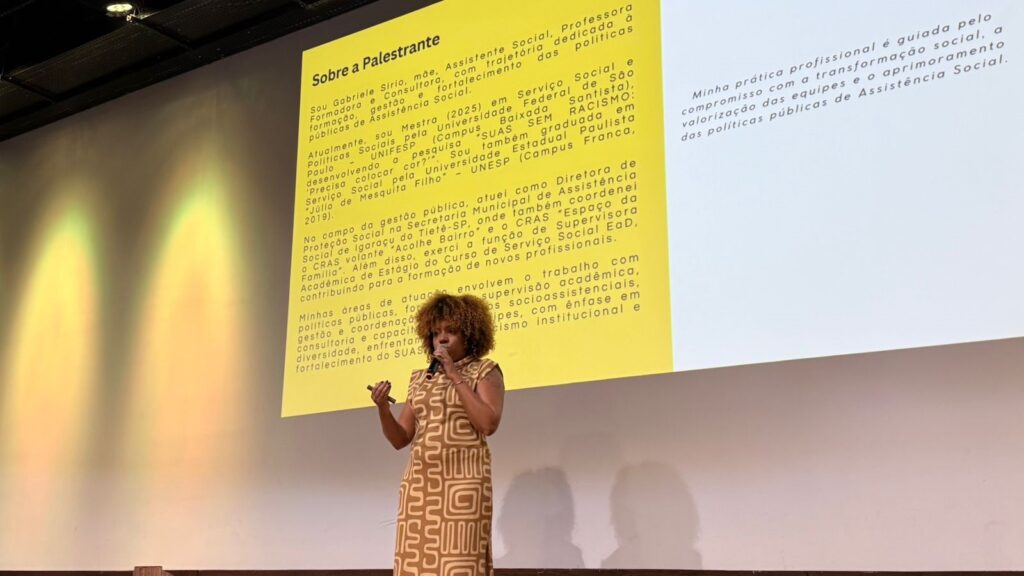 A imagem mostra uma mulher palestrante chamada Gabriele Sirio, que está falando em um evento ou conferência. Ela está posicionada à frente de um microfone e segura um objeto na mão, possivelmente um controle para apresentação. Atrás dela, há uma grande tela projetando um slide com texto em português que apresenta seu currículo e trajetória profissional. O slide tem um fundo amarelo claro com texto em preto e branco. O título principal é "Sobre a Palestrante". Gabriele se apresenta como mãe, assistente social, professora formadora e consultora, com experiência em formação, gestão e fortalecimento de políticas públicas de Assistência Social. Ela menciona que está cursando mestrado em Serviço Social e Políticas Sociais na Universidade Federal de São Paulo, com uma pesquisa sobre racismo no Sistema Único de Assistência Social (SUAS). Também é graduada em Serviço Social pela Universidade Estadual Paulista. No texto, detalha sua atuação na gestão pública, incluindo cargos como Diretora de Proteção Social na Secretaria Municipal de Assistência Social de Igaraçu do Tietê-SP e coordenação de unidades do CRAS (Centro de Referência de Assistência Social). Além disso, atua como supervisora acadêmica de estágio no curso de Serviço Social EaD. Sua prática profissional é guiada pelo compromisso com a transformação social, valorização das equipes e aprimoramento das políticas públicas de Assistência Social. A imagem transmite um contexto de palestra ou aula voltada para temas sociais e de políticas públicas.