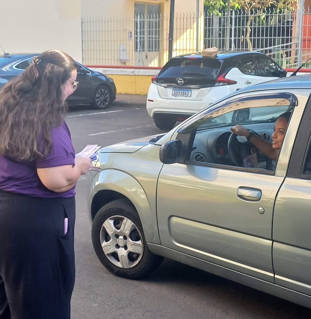 A imagem mostra duas mulheres em um estacionamento. Uma mulher, vista de costas, está em pé do lado esquerdo da imagem, vestindo uma blusa roxa e calças pretas. Ela tem cabelo castanho longo e ondulado e usa óculos. Ela está segurando alguns papéis nas mãos. A outra mulher está sentada no banco do motorista de um carro prateado claro à direita. Ela tem pele mais escura, cabelo escuro e está sorrindo enquanto olha para a mulher em pé. Ela está com a mão direita no volante. Atrás do carro prateado, há um carro branco com a placa "SSV3D62". Há também um carro escuro estacionado mais atrás à esquerda. O estacionamento parece ser ao ar livre, com edifícios e uma cerca visíveis ao fundo. A luz do sol sugere que é dia.