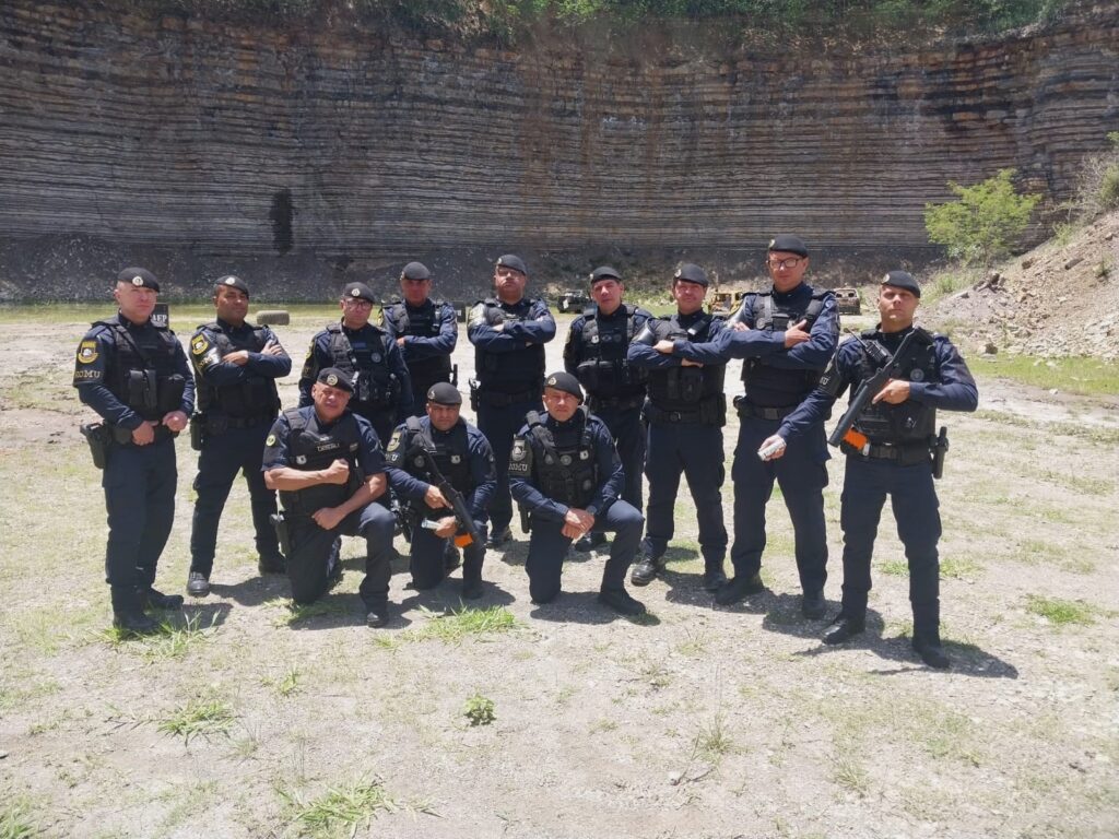 A imagem mostra um grupo de 11 homens uniformizados, vestindo calças azuis escuras, camisas azuis escuras com coletes táticos pretos, boinas pretas e botas pretas. Eles parecem ser policiais ou membros de uma força de segurança, pois alguns usam distintivos em seus coletes e cintos com equipamentos. O grupo está posicionado em uma área externa com um fundo de uma parede de rocha com camadas horizontais visíveis, sugerindo uma pedreira ou uma formação geológica natural. O chão em primeiro plano é coberto por terra e grama esparsa. A maioria dos homens está em pé, com alguns cruzados de braços, enquanto outros estão ajoelhados ou agachados. Um deles, à direita, está segurando uma arma longa, e outro, mais à esquerda, parece estar segurando uma arma curta em seu coldre. A iluminação sugere que a foto foi tirada durante o dia.