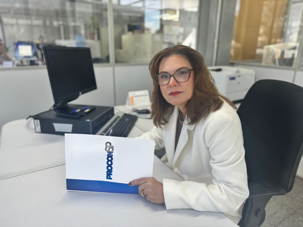 A imagem mostra uma mulher sentada em uma mesa de escritório, com um computador e uma pasta com o logo "PROCONSP" à sua frente. Ela usa um blazer branco e óculos, e olha diretamente para a câmera com uma expressão séria. O ambiente parece ser um escritório moderno, com divisórias de vidro e outros equipamentos de escritório ao fundo.