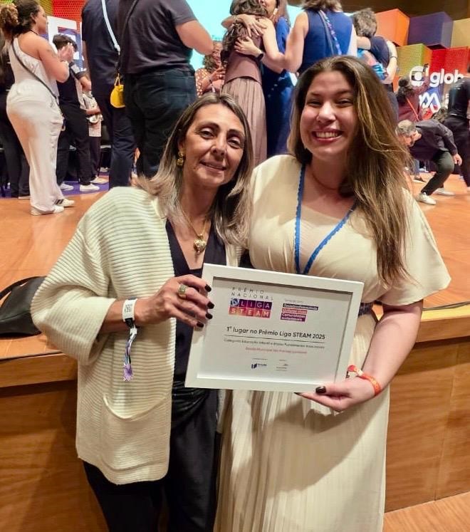 A imagem mostra duas mulheres sorrindo para a câmera, segurando um certificado. A mulher à esquerda, com cabelo castanho e ondulado, usa um cardigã bege sobre uma blusa escura e um colar dourado. Ela usa um anel grande em seu dedo indicador. A mulher à direita, com cabelo castanho longo e liso, usa um vestido de cor creme com detalhes em azul. Ela tem uma pulseira laranja no pulso esquerdo. O certificado que elas seguram tem o título "PREMIO NACIONAL LIGA STEAM 2025" e indica "1º lugar no Prêmio Liga STEAM 2025". Ao fundo, há outras pessoas em um ambiente que parece ser um evento ou premiação, com um palco e um logo em forma de globo ao fundo.