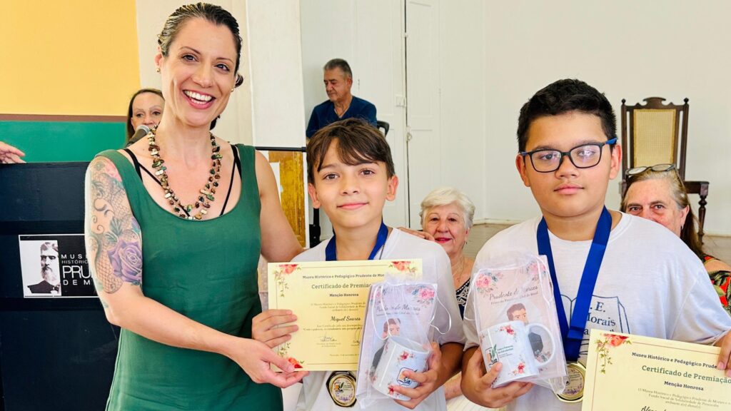 Na imagem, uma mulher sorridente, com um vestido verde e um colar de contas coloridas, está em pé à esquerda. Ela tem uma tatuagem em seu braço esquerdo. Ela segura um certificado e presente para dois meninos que estão à sua direita. Os meninos, um com óculos e outro sem, também estão sorrindo e usam medalhas no pescoço. Um deles segura um presente embrulhado com um laço rosa e uma caneca. Ao fundo, outras pessoas estão presentes, incluindo um homem mais velho sentado e uma mulher mais velha em pé atrás dos meninos. O ambiente parece ser uma cerimônia de premiação, possivelmente em um museu ou instituição educacional, como sugerido pelos certificados e pelo homem retratado em um cartaz à esquerda.