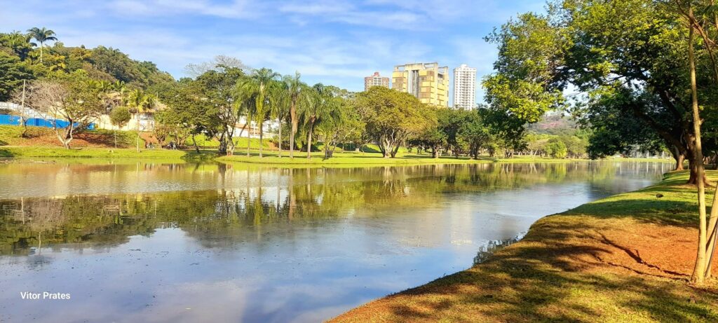 A imagem exibe uma paisagem serena de um lago em um parque, com reflexos nítidos das árvores e do céu azul na água calma. Ao fundo, uma colina verdejante serve de pano de fundo para um conjunto de edifícios modernos, contrastando com a natureza exuberante. As árvores frondosas, incluindo palmeiras, margeiam a água, criando um ambiente convidativo para o lazer. A grama verde e bem cuidada, com manchas de terra avermelhada, compõe a margem do lago, onde a luz do sol cria sombras projetadas pelas árvores. A cena é complementada por um céu azul claro com algumas nuvens brancas, sugerindo um dia agradável. Ao canto inferior esquerdo, encontra-se a assinatura "Vitor Prates".
