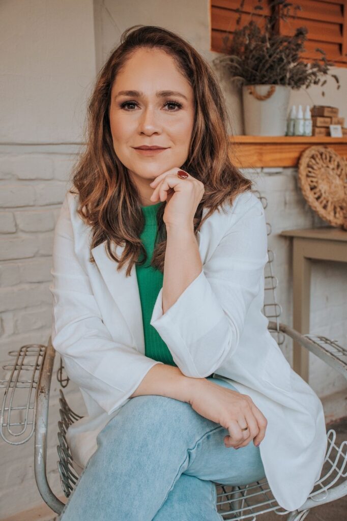 A mulher está sentada em uma cadeira de metal com um blazer branco aberto sobre uma blusa verde. Ela usa jeans claros e tem uma mão apoiada no queixo, com um anel vermelho na mão. A outra mão está descansando no colo. O cabelo dela é castanho e ondulado, e ela tem uma maquiagem suave. Ao fundo, há uma parede de tijolos brancos, uma prateleira com plantas e alguns frascos, e uma cesta de palha. A iluminação é suave e natural.