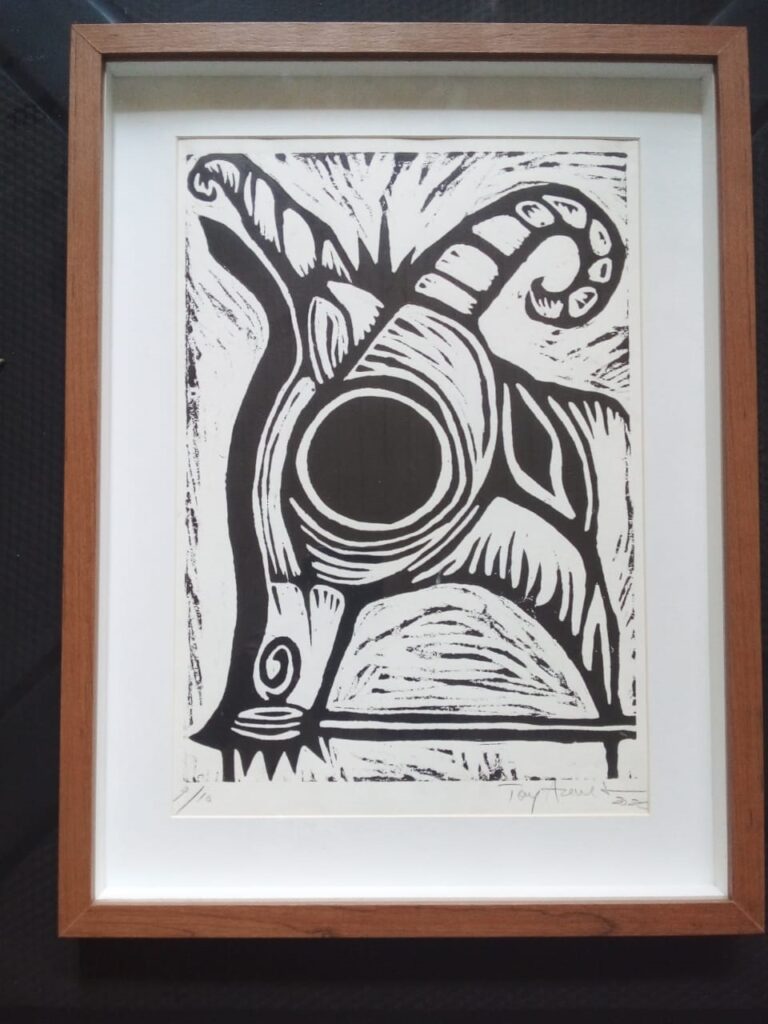 A imagem exibe uma gravura em preto e branco, emoldurada com uma moldura de madeira clara. A gravura, que parece ser uma xilogravura ou linoleogravura, apresenta uma representação estilizada de um bode ou carneiro. O animal é retratado de forma abstrata e expressiva, com linhas fortes e contrastantes. A cabeça do bode domina a composição, com um grande chifre em espiral que se curva para cima e para a direita. O olho é representado por um círculo escuro e proeminente, cercado por padrões concêntricos que sugerem um efeito visual dinâmico. O corpo do animal é formado por formas geométricas e texturas que transmitem movimento e energia. A técnica utilizada na gravura é evidente nas áreas de hachuras e nas texturas criadas pelas linhas, que dão profundidade e volume à imagem. Na parte inferior da gravura, há uma assinatura do artista e a data "22", além de uma numeração em algarismos arábicos, "1/15", que indica que esta é a primeira cópia de uma tiragem limitada de quinze exemplares. A gravura está montada sobre um passe-partout branco, que realça a obra e a protege. A moldura de madeira clara, com um tom amadeirado suave, complementa a estética da gravura, conferindo-lhe um ar de elegância e simplicidade.