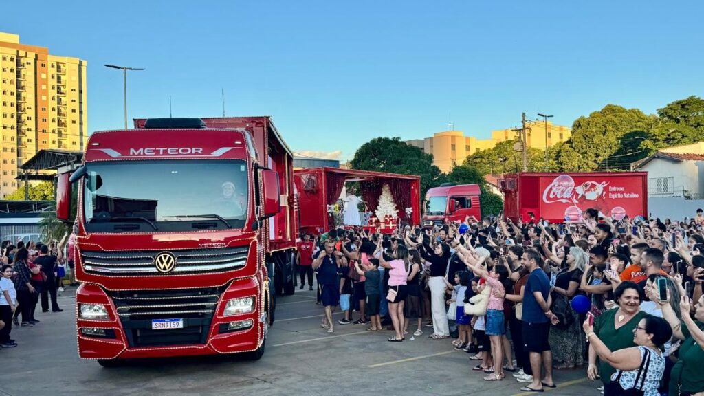 O caminhão da frente, um Volkswagen Meteor vermelho, está adornado com luzes de Natal brancas ao longo de sua carroceria e grade. No para-brisa, é possível ver uma pessoa sorrindo, possivelmente o motorista, que também usa um gorro de Papai Noel. A placa do caminhão exibe "SRN9159". Atrás deste, há outro caminhão vermelho com uma plataforma elevada, que serve como um palco. Sobre esta plataforma, há uma árvore de Natal branca decorada, presentes e uma pessoa vestida de branco, talvez representando um anjo ou a figura do Natal. Mais atrás, um terceiro caminhão vermelho ostenta o icônico logo da Coca-Cola e a frase "Entre no Espírito Natalino", reforçando a temática festiva e a associação da marca com as celebrações de fim de ano. Uma grande multidão de pessoas se aglomera em frente aos caminhões, muitas delas com os braços erguidos, segurando celulares para tirar fotos e gravar vídeos. Há uma atmosfera de celebração e expectativa, com pessoas de todas as idades aproveitando o evento. O céu azul e a luz do sol sugerem que a carreata está acontecendo durante o dia, possivelmente no final da tarde.