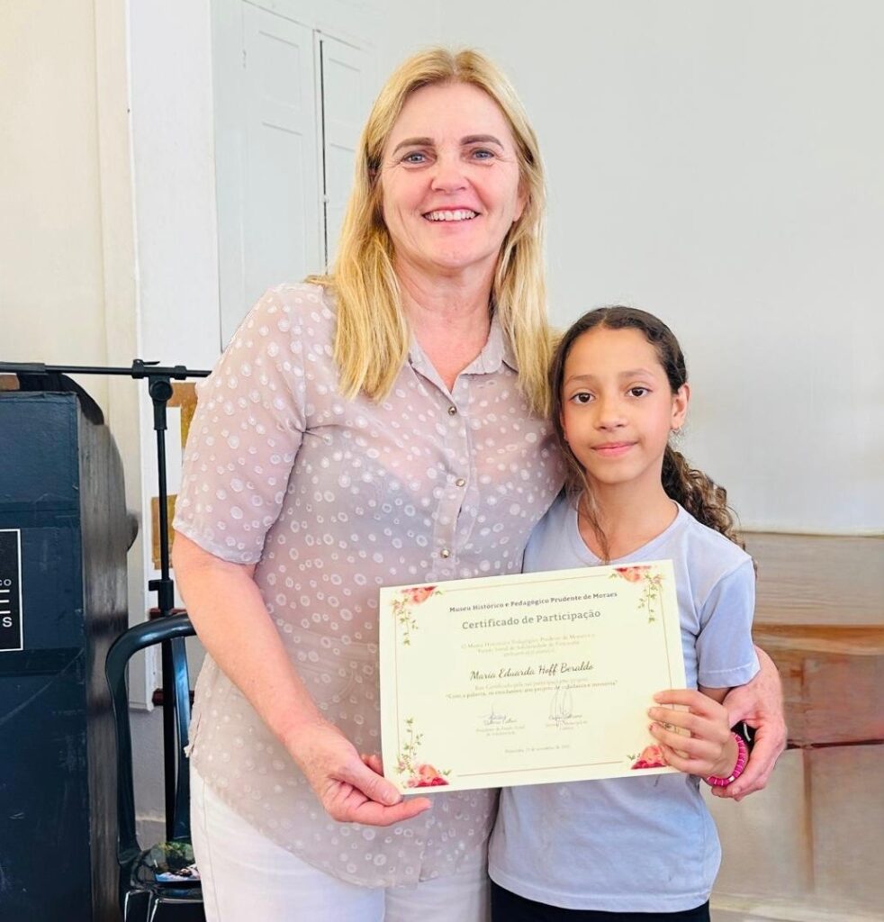 Em uma sala com paredes claras e uma porta branca ao fundo, uma mulher de cabelos loiros e pele clara, sorrindo, abraça uma menina de cabelos castanhos. A mulher veste uma blusa clara com bolinhas em tom mais escuro e calça clara. A menina veste uma blusa cinza clara. Ambas seguram um certificado com a mão direita da mulher e a mão esquerda da menina. O certificado, com bordas decoradas com flores rosas, tem o título "Certificado de Participação" e o nome "Maria Eduarda Hoff Beraldo" em destaque. Ao fundo, à esquerda, há um objeto escuro com detalhes metálicos, possivelmente um piano ou um armário antigo. À direita, um banco de madeira.