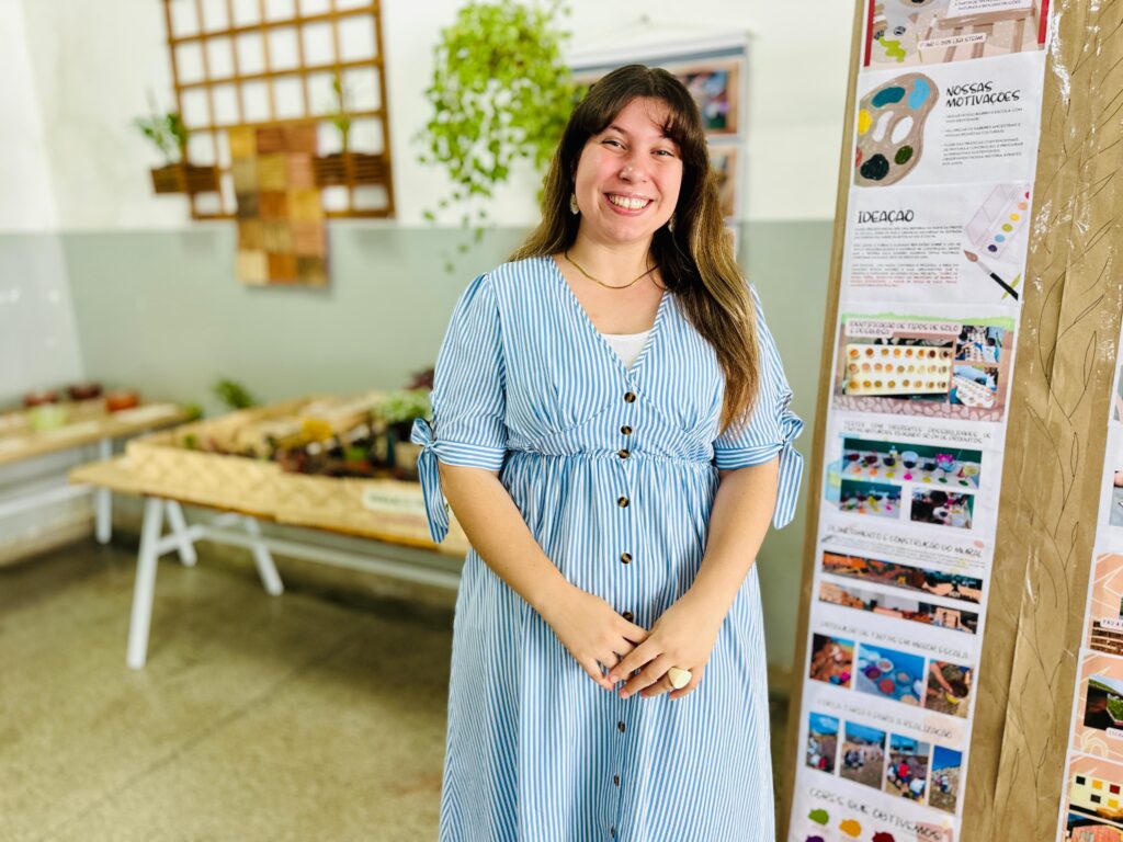 A mulher sorri para a câmera em um vestido listrado azul e branco. Ela está em pé em uma sala com exposições em mesas e em um painel de exibição. O painel tem textos e imagens que parecem ser de um projeto escolar ou acadêmico. Há plantas penduradas no fundo, e um rack de madeira com vasos de plantas na parede. As mesas atrás dela exibem maquetes ou displays de projetos, possivelmente relacionados a ciências ou artes.