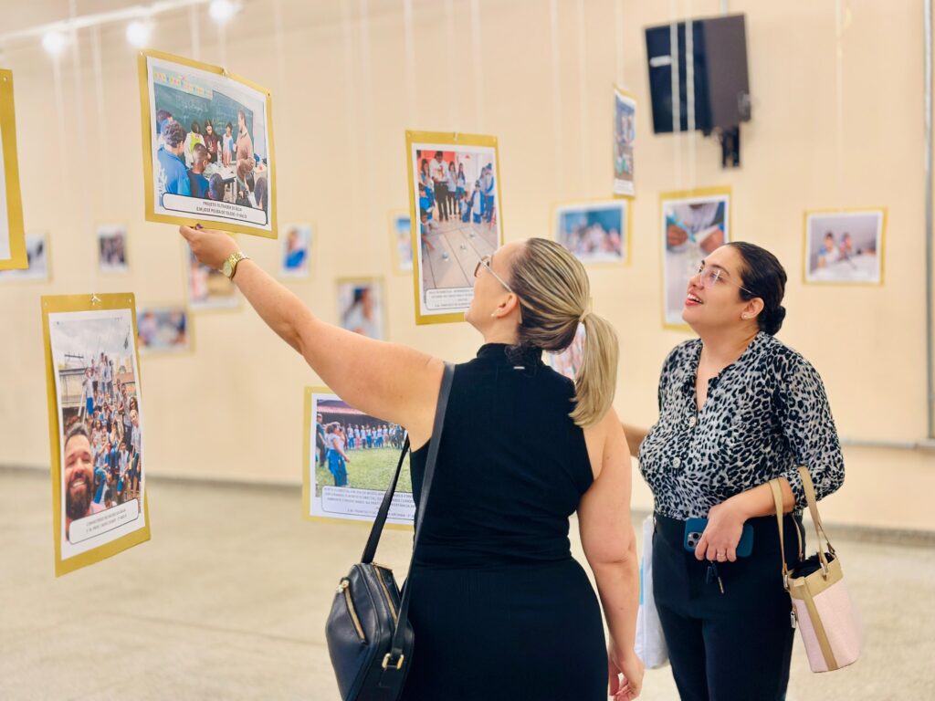Duas mulheres estão em uma galeria de arte, olhando para uma exposição de fotografias suspensas. A mulher da esquerda, com um rabo de cavalo loiro, usa um vestido preto sem mangas e tem uma bolsa preta a tiracolo. Ela está estendendo a mão para tocar uma das fotos. A mulher da direita, com cabelo escuro preso em um coque, usa uma blusa estampada de leopardo e calças pretas. Ela está segurando um celular e um chaveiro na mão direita, e uma bolsa bege e rosa na mão esquerda. Ambas as mulheres parecem absortas na exposição. As fotos são penduradas em fios finos e parecem ser de eventos escolares ou comunitários, com muitas pessoas em cada imagem. O fundo é uma parede de cor clara com algumas luminárias e um alto-falante pendurado.