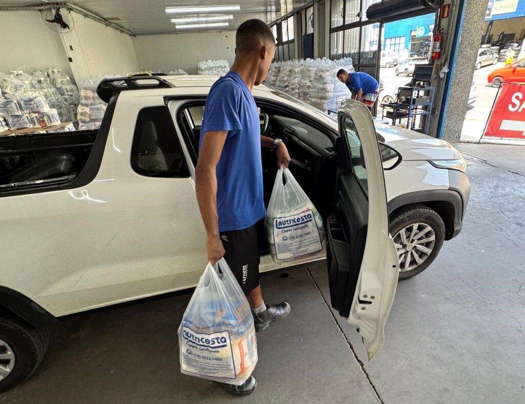 A imagem mostra um jovem vestindo uma camiseta azul e shorts pretos, carregando duas sacolas plásticas com compras. Ele está em pé ao lado de uma caminhonete branca com a porta do passageiro aberta. As sacolas plásticas são transparentes e contêm o logotipo "nutricesta Compra Inteligente" e um número de telefone. Uma das sacolas está no chão, perto dos pés do jovem, enquanto a outra está em sua mão direita. Ao fundo, é possível ver um galpão com prateleiras cheias de sacos brancos empilhados, possivelmente alimentos ou suprimentos. Há também um outro homem de camiseta azul e shorts vermelhos, agachado, mexendo em algo em uma área de carregamento. A cena sugere uma operação de entrega ou recebimento de mercadorias em um local de armazenamento ou comércio. A iluminação é forte, indicando um ambiente externo ou bem iluminado.
