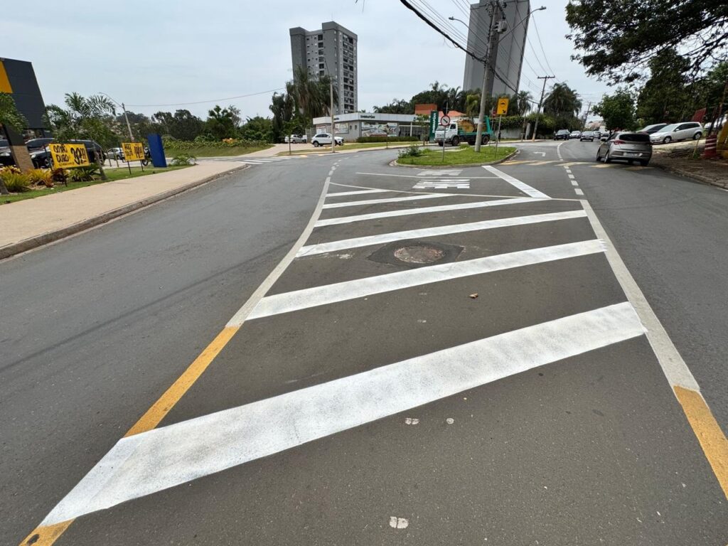 A imagem mostra uma rua com uma faixa de pedestres pintada em branco e amarelo. A faixa de pedestres está localizada em uma curva na estrada, com várias faixas de tráfego. Há carros estacionados e em movimento na estrada, bem como edifícios e árvores ao fundo. O céu está nublado.