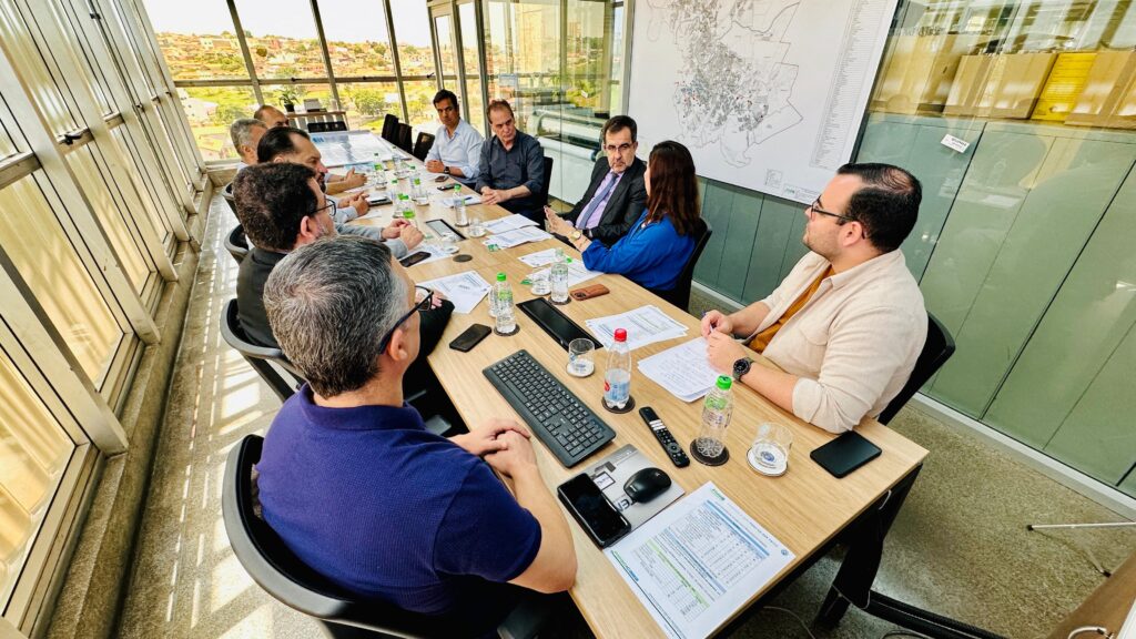 A imagem mostra um grupo de pessoas reunidas em uma sala de reuniões moderna, com grandes janelas que oferecem uma vista panorâmica de uma cidade e montanhas ao fundo. As janelas também permitem a entrada de muita luz natural, criando um ambiente claro e arejado. No centro da sala, há uma longa mesa de madeira onde os participantes estão sentados, engajados em uma discussão. Sobre a mesa, há vários documentos, garrafas de água, copos, um teclado, um mouse, um controle remoto e smartphones. A disposição dos objetos sugere que a reunião está em andamento, com materiais de apoio sendo utilizados. Os participantes, em sua maioria homens vestidos de forma casual de negócios, parecem atentos e envolvidos na conversa. Uma mulher, vestida de azul, está falando, gesticulando com as mãos, enquanto um homem de terno e gravata à sua frente a escuta atentamente. No geral, a cena transmite uma atmosfera de profissionalismo e colaboração.