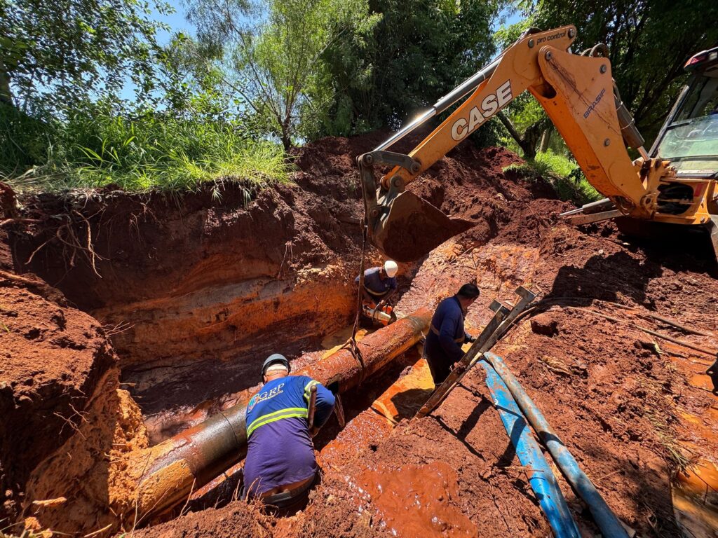 A imagem mostra uma equipe trabalhando na substituição de uma tubulação de água em uma área externa, possivelmente em uma construção ou manutenção de infraestrutura. * *Trabalhadores:* Três homens vestidos com uniformes de trabalho azuis, capacetes e botas estão envolvidos na tarefa. Dois deles estão focados em conectar uma nova seção de tubulação a uma válvula azul grande e a uma seção existente da tubulação. O terceiro homem, com óculos, observa o trabalho. * *Equipamentos:* Uma escavadeira laranja com a caçamba levantada paira sobre a área, sugerindo que a tubulação está sendo manuseada ou posicionada com auxílio mecânico. Ferramentas como chaves de boca estão visíveis em baldes amarelos e vermelhos, indicando o trabalho manual envolvido. * *Ambiente:* O local é lamacento e coberto de terra vermelha, com pilhas de terra ao redor, o que sugere que a área foi escavada para acessar a tubulação. Árvores e vegetação ao fundo indicam um ambiente ao ar livre. A cena transmite a ideia de um trabalho árduo e essencial para a manutenção de serviços públicos.