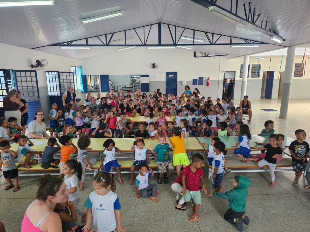 A imagem mostra um grupo grande de crianças, a maioria em idade escolar, reunidas em um salão com mesas e bancos. Há também adultos presentes, aparentando ser professores ou cuidadores. As crianças estão vestidas com roupas coloridas e algumas usam uniformes escolares. O ambiente parece ser uma escola ou um centro comunitário, com janelas e portas ao fundo, além de um ventilador de teto e luminárias. Algumas crianças estão sentadas nas mesas, outras em pé ou brincando no chão. A atmosfera geral é alegre e descontraída, sugerindo um evento ou atividade escolar.
