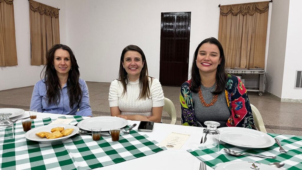 A imagem mostra três mulheres sentadas em uma mesa longa, com uma toalha xadrez verde e branca. Elas estão em um ambiente interno, possivelmente um salão de eventos ou restaurante, com cortinas marrons ao fundo e uma porta escura entre as duas primeiras mulheres. A mulher à esquerda, com cabelo escuro e liso, veste uma camisa azul clara e sorri para a câmera. Na mesa à sua frente, há um prato com alguns pastéis fritos, um copo pequeno com líquido marrom e talheres. A mulher do meio, com cabelo castanho e ondulado, usa uma blusa branca de gola alta com um padrão texturizado e também sorri. Em sua frente, há um prato branco, um celular e um menu. A mulher à direita, com cabelo escuro e solto, veste uma blusa cinza e um casaco colorido com estampas vibrantes. Ela usa um colar de contas laranjas e sorri amplamente. Na mesa, há um prato, um copo de vidro e talheres. Ao fundo, percebe-se um carrinho de serviço com bandejas e uma grade de ventilação na parede. O ambiente parece ser um local formal, possivelmente para um almoço ou jantar.