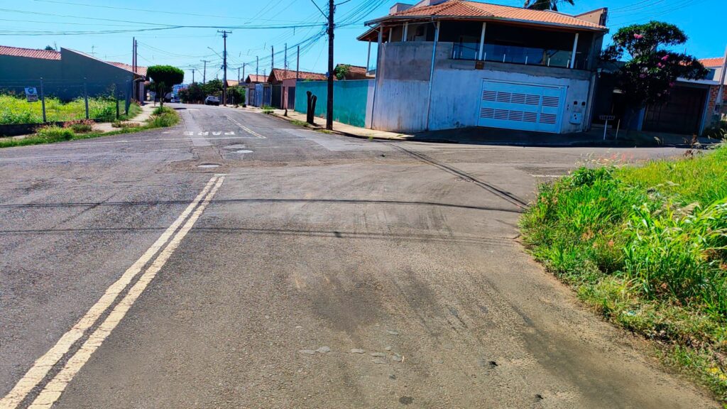 A imagem mostra uma rua residencial em um dia ensolarado, vista em um ângulo ao nível dos olhos e em plano médio. A rua de asfalto ocupa a parte inferior da imagem, com linhas amarelas duplas no centro. À direita, há um gramado verde com algumas flores pequenas e altas. Ao fundo, há casas de ambos os lados da rua, incluindo uma casa moderna de dois andares com fachada cinza, garagem branca e uma varanda com vidro. O céu está azul e limpo, e postes de energia com muitos fios elétricos estão alinhados ao longo da rua, evidenciando uma área urbana. A iluminação é forte, criando sombras e destacando texturas. No chão, está pintada a palavra "PARE", indicando uma sinalização de trânsito. A cena transmite a sensação de um bairro residencial tranquilo em um dia claro e ensolarado.