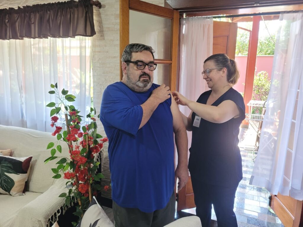 A imagem mostra um homem de meia-idade, com barba e óculos, vestindo uma camiseta azul, recebendo uma vacina no braço. Uma mulher, provavelmente uma enfermeira, vestindo uniforme escuro e óculos, está aplicando a injeção. Ela tem um crachá preso à roupa. O ambiente parece ser uma sala de estar ou um consultório médico domiciliar, com uma poltrona branca à esquerda, decorada com almofadas com estampas botânicas. Há também um arranjo de flores artificiais vermelhas em um vaso. Ao fundo, cortinas brancas cobrem janelas, e uma porta de madeira está entreaberta, revelando um ambiente externo ensolarado. O chão é revestido com um padrão de ladrilhos.