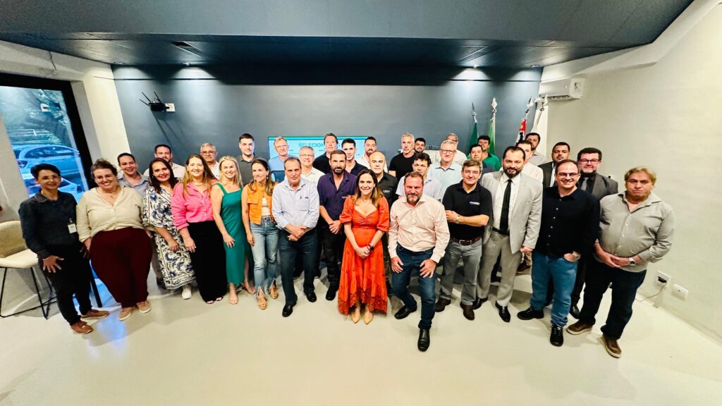 A imagem mostra um grupo grande de pessoas, cerca de 34 indivíduos, reunidos em um ambiente interno que parece ser um escritório ou uma sala de reuniões. Eles estão organizados em várias fileiras, com alguns em pé na frente e outros atrás, formando uma composição em camadas. A maioria está olhando para a câmera, com expressões neutras ou leves sorrisos, transmitindo uma atmosfera profissional e amigável. Ao fundo, há uma tela grande com a palavra "SELECAO" escrita em letras brancas, sugerindo que o encontro pode estar relacionado a um processo de seleção, reunião ou evento corporativo. Também é possível ver algumas bandeiras à direita da imagem, o que adiciona um tom formal e institucional ao ambiente. O local possui paredes em tons escuros, provavelmente azul e cinza, e o piso é claro. A iluminação é artificial, destacando bem as pessoas e o espaço. À esquerda, há uma janela que mostra parte do exterior, incluindo árvores e um carro estacionado, indicando que o ambiente está em um prédio com vista para a rua. As roupas dos participantes variam entre o casual e o formal, com homens usando camisas e algumas gravatas, e mulheres vestindo blusas, vestidos e calças, o que reforça a ideia de um contexto profissional. No geral, a imagem transmite a sensação de uma reunião organizada, possivelmente de uma equipe de trabalho, grupo de estudo, ou evento relacionado a seleção ou apresentação institucional.