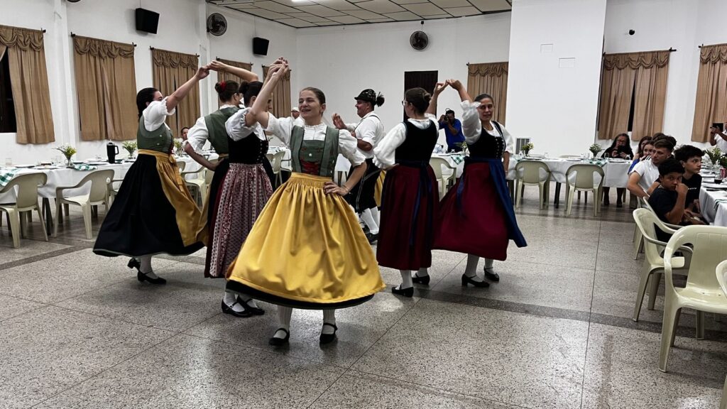 A imagem mostra um grupo de jovens dançando em um salão. Eles estão vestidos com trajes tradicionais, que parecem ser de origem europeia, com saias longas e volumosas, blusas brancas e coletes. As cores predominantes nos trajes são o amarelo, o verde, o bordô e o preto. As dançarinas estão de mãos dadas, formando um círculo e se movendo em sincronia, com expressões alegres nos rostos. Ao fundo, mesas estão postas com toalhas brancas e decorações florais, sugerindo que o evento é uma celebração ou festa. Algumas pessoas estão sentadas às mesas, observando a dança. O ambiente é iluminado e parece ser um espaço interno, possivelmente um salão de festas ou um centro comunitário.