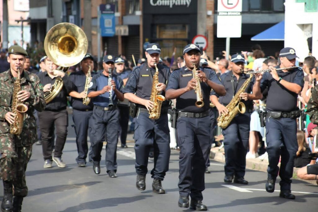 A imagem mostra uma banda militar desfilando em uma rua. Vários músicos estão tocando instrumentos de sopro, incluindo saxofones, um clarinete e um trombone. Eles estão vestindo uniformes militares e de polícia. Ao fundo, há pessoas assistindo ao desfile e edifícios. A atmosfera parece ser de celebração ou evento cívico.