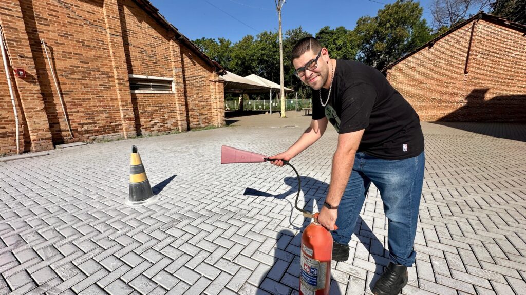 A imagem mostra um homem sorridente, com óculos e barba, usando uma camiseta preta e jeans. Ele está agachado e segurando um extintor de incêndio vermelho na mão direita, enquanto com a mão esquerda aponta para um cone de trânsito preto e amarelo. O extintor está conectado a um bico rosa, que ele segura. O cenário é um pátio pavimentado com blocos de concreto dispostos em um padrão de espinha de peixe. Ao fundo, há um prédio de tijolos vermelhos e algumas árvores sob um céu azul claro. A iluminação sugere que é um dia ensolarado, com sombras projetadas no chão.