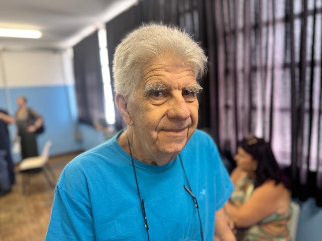 A imagem em close-up mostra um homem idoso com cabelos brancos e pele enrugada, olhando para a câmera com um leve sorriso. Ele está vestindo uma camiseta azul clara e tem óculos pendurados em seu pescoço. O fundo está desfocado, mas parece ser um ambiente interno, possivelmente uma sala de reuniões ou um salão de eventos, com outras pessoas e cortinas escuras visíveis.