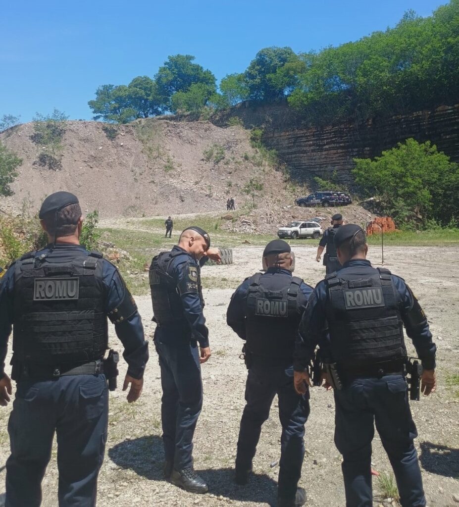 A imagem mostra um grupo de policiais em um ambiente externo, possivelmente uma pedreira ou área de treinamento. Quatro policiais estão em primeiro plano, de costas para a câmera, vestindo uniformes escuros com coletes táticos que exibem a sigla "ROMU". Eles usam boinas pretas. Ao fundo, o terreno é irregular, com uma grande pilha de terra e rochas à esquerda e uma formação rochosa em camadas à direita. Há também algumas árvores e vegetação espalhadas pela paisagem. No centro e à direita do plano de fundo, alguns veículos estão estacionados, incluindo um carro branco e um carro escuro. Várias outras pessoas, possivelmente policiais ou pessoal de apoio, estão visíveis em segundo plano, perto dos veículos e na encosta. O céu está azul e claro, indicando um dia ensolarado.