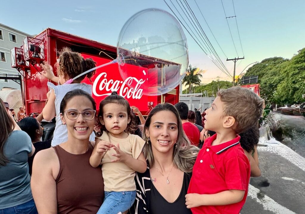 A imagem mostra um grupo de pessoas em frente a um caminhão vermelho da Coca-Cola. No centro da imagem, duas mulheres e duas crianças estão posando para a foto. A mulher à esquerda, usando óculos e uma blusa marrom, segura uma menina pequena no colo. A menina, com roupas claras, olha para a câmera com uma expressão séria. A mulher à direita, com cabelos longos e uma blusa preta, sorri para a câmera. Ao lado dela, um menino pequeno, vestindo uma camisa polo vermelha, olha para o lado com curiosidade. Ao fundo, o caminhão da Coca-Cola é o destaque, com o logotipo da marca em destaque. Uma grande bolha transparente está suspensa no ar, adicionando um elemento lúdico à cena. Outras pessoas podem ser vistas ao fundo, sugerindo um evento ou celebração. O céu ao entardecer, com tons alaranjados e azuis, contribui para a atmosfera festiva da imagem. A presença de árvores e postes de eletricidade indica que o evento está ocorrendo em uma área urbana ou suburbana.