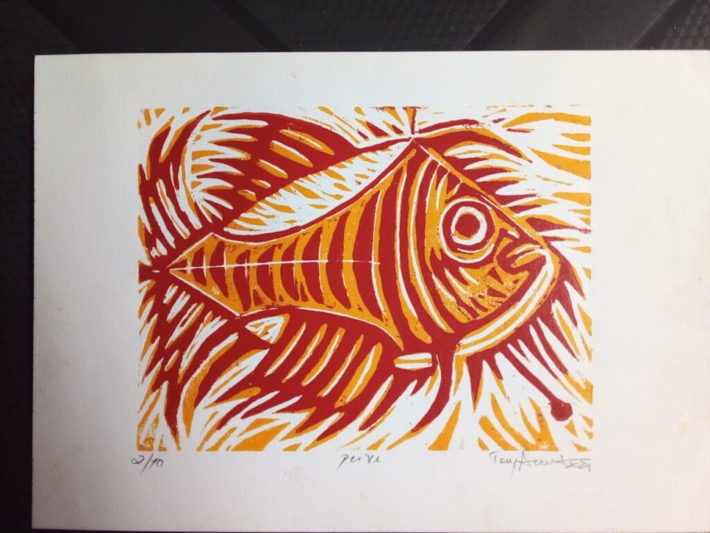 A imagem é uma gravura em linóleo de um peixe estilizado em tons de vermelho e amarelo, com o fundo em branco. O peixe está representado de perfil, com escamas marcadas por linhas paralelas e um olho circular proeminente. A técnica de gravura é evidente nas linhas fortes e na textura criada pela sobreposição das cores. No canto inferior esquerdo da imagem, há uma numeração escrita à mão: "2/10", indicando que esta é a segunda cópia de uma edição limitada de dez. Ao lado, centralizado, está a palavra "peixe" escrita em letras minúsculas. No canto inferior direito, encontra-se a assinatura do artista, "Tony Arut", seguida pelo ano "2024". A composição geral é vibrante e expressiva, com o movimento sugerido pelas linhas onduladas do fundo que parecem imitar a água.