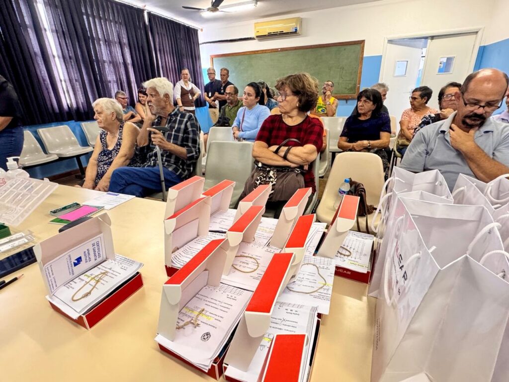 A imagem mostra um grupo de pessoas, predominantemente idosos, sentadas em cadeiras em uma sala. Na frente da sala, sobre uma mesa, há várias caixas vermelhas e brancas abertas, contendo certificados. Há também sacolas brancas de presente e outros documentos espalhados pela mesa. As pessoas na plateia parecem atentas, com algumas olhando para a frente, onde provavelmente está o orador ou o objeto da apresentação. O ambiente sugere um evento comunitário, uma palestra ou a entrega de algum tipo de benefício.