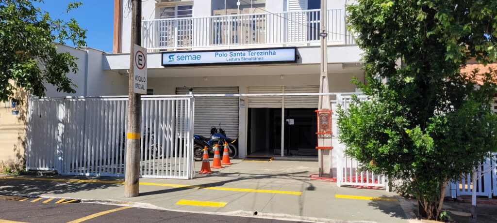 A imagem mostra a fachada de um prédio com uma placa luminosa que diz "semae Polo Santa Terezinha Leitura Simultânea". Há um portão branco de metal aberto, revelando uma entrada com uma porta de rolo. Na frente do portão, há cones de trânsito laranja e duas motocicletas. Um poste de luz está à esquerda da entrada, com uma placa de trânsito circular com a letra "E" dentro e a inscrição "VEÍCULO OFICIAL" abaixo. Uma árvore frondosa com folhas verdes cresce à direita da entrada. O céu está azul e ensolarado.