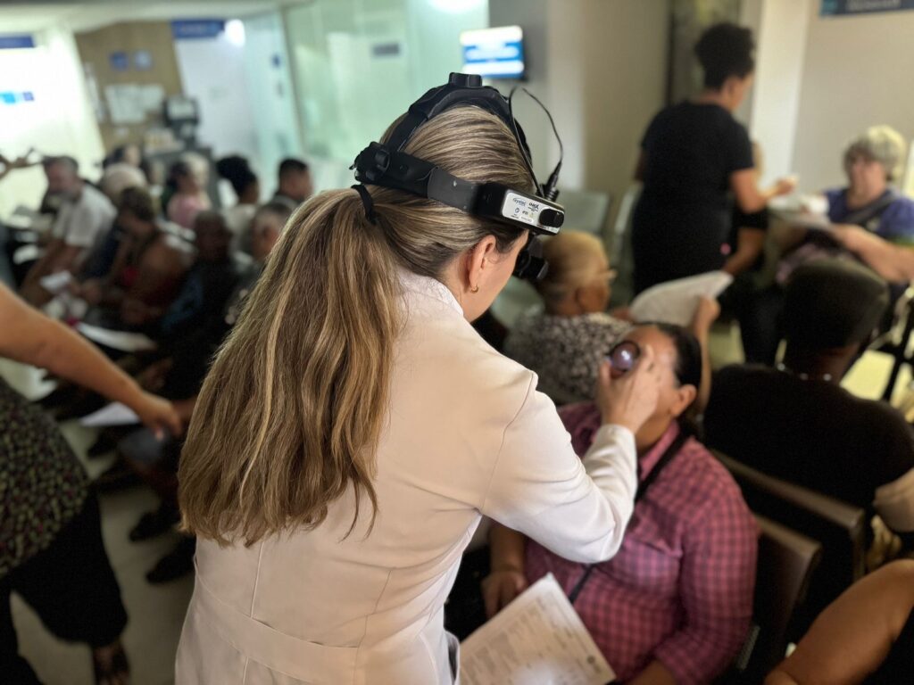 A imagem mostra uma profissional de saúde, vestida com um jaleco branco e usando um oftalmoscópio de cabeça, examinando o olho de uma paciente. A paciente, que está sentada, usa uma camisa xadrez rosa e tem a mão levantada para auxiliar no exame. Ao fundo, várias outras pessoas estão sentadas em cadeiras, aguardando atendimento em uma sala de espera que parece ser de uma clínica ou hospital, com sinalizações e móveis típicos desse ambiente. A iluminação sugere um ambiente interno, possivelmente durante o dia.