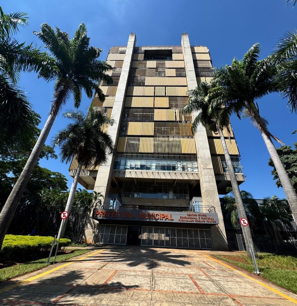 A imagem mostra a fachada de um edifício público, provavelmente a prefeitura de uma cidade, com uma arquitetura moderna e imponente. O edifício é alto e estreito, com uma estrutura de concreto aparente e uma fachada com painéis em tons de amarelo e bege, que criam um padrão reticulado. Palmeiras altas e frondosas emolduram o edifício em primeiro plano, adicionando um toque tropical à cena. O céu azul e claro, juntamente com a iluminação intensa, sugere um dia ensolarado. Na base do edifício, há uma entrada com portões metálicos fechados e uma placa com o nome "PREFEITURA MUNICIPAL" em letras grandes e alaranjadas. Abaixo, há outra placa menor com os dizeres "CENTRO CÍVICO, EDUCACIONAL E CULTURAL FLORIVALDO COELHO PRATES". Sinais de trânsito circulares com a letra "E" dentro de um círculo vermelho, indicando "Estacionamento Proibido", estão posicionados em ambos os lados da entrada. O caminho em frente ao edifício é pavimentado com pedras e delimitado por faixas amarelas, com grama verde e árvores ao redor.
