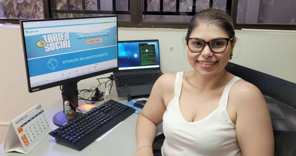 A imagem mostra uma mulher sorrindo para a câmera, usando óculos e uma regata branca. Ela está sentada em uma mesa de escritório com dois monitores de computador. O monitor da esquerda exibe um site com o título "TARIFA SOCIAL" e informações sobre "SITUAÇÃO DO ABASTECIMENTO". O monitor da direita, um laptop, exibe uma tela com código em verde. Ao lado dos monitores, há um teclado, um mouse e um calendário de mesa que marca "NOVEMBRO 2025". Há também alguns objetos decorativos e materiais de escritório na mesa. A iluminação sugere que a foto foi tirada em um ambiente interno, possivelmente um escritório.
