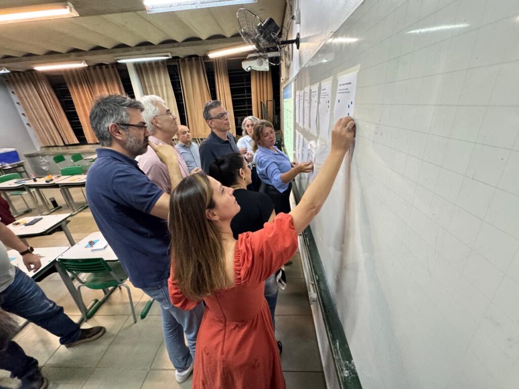 A imagem mostra um grupo de adultos em uma sala que parece ser uma sala de aula ou de reunião. Eles estão interagindo com um quadro branco onde estão fixadas várias folhas com pontos coloridos e textos. No centro da imagem, uma mulher vestindo um vestido laranja está de braços levantados, tocando ou apontando para uma dessas folhas no quadro. Ao lado dela, outra mulher, vestida com uma blusa azul clara, também está focada nas folhas do quadro. Ao fundo, outras pessoas observam ou conversam entre si. A sala está iluminada por luzes fluorescentes no teto e possui mesas e cadeiras organizadas, típicas de um ambiente educacional ou de oficina. A atmosfera sugere uma atividade colaborativa ou uma dinâmica de grupo, com as pessoas envolvidas e concentradas na tarefa.