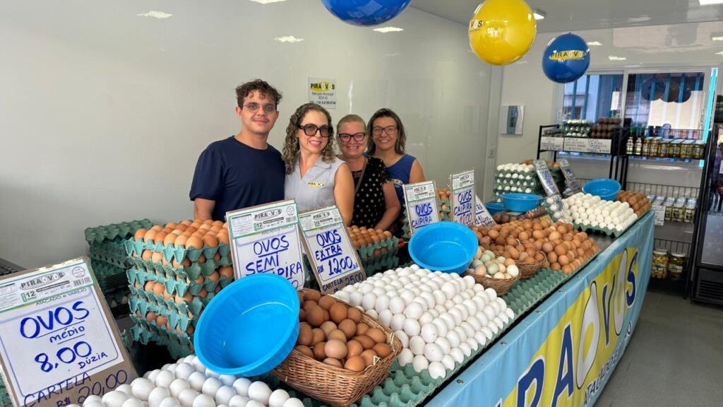 A imagem mostra uma loja de ovos com quatro pessoas atrás do balcão, sorrindo para a câmera. O balcão está repleto de caixas e cestos de ovos de diferentes tamanhos e cores, incluindo ovos brancos, marrons e caipiras. Há também placas com os preços dos ovos, como "OVOS MÉDIO 8,00 DUZIA" e "OVOS EXTRA R$ 12,00 CARTELA R$ 25,00". Ao fundo, prateleiras exibem mais produtos, como garrafas e potes. Balões azuis e amarelos com o logo da loja "PIRA OVOS" estão pendurados no teto.