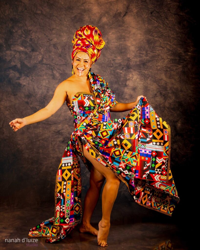 A imagem mostra uma mulher sorrindo em frente a um fundo escuro texturizado. Ela está vestindo um vibrante traje africano com um turbante combinando. O vestido é feito de um tecido colorido com um padrão geométrico ousado em tons de vermelho, amarelo, azul, verde, preto e branco. O vestido é envolto em torno de seu corpo, com uma abertura alta que revela suas pernas. O turbante é alto e volumoso, com dobras de tecido em vermelho, amarelo e roxo. O rosto da mulher está adornado com pintura facial branca, incluindo linhas que descem de seus olhos e em suas bochechas. Ela tem um sorriso largo e parece estar em movimento, com um braço estendido e o outro segurando a saia do vestido. Seus pés estão descalços, com as pontas dos pés levantadas, sugerindo uma pose de dança. A iluminação na foto é dramática, com a mulher bem iluminada e o fundo escurecendo gradualmente. A marca d'água "nanah d'luize studio" aparece no canto inferior esquerdo da imagem. O estilo geral da fotografia é vibrante e enérgico, celebrando a cultura e a beleza do traje.