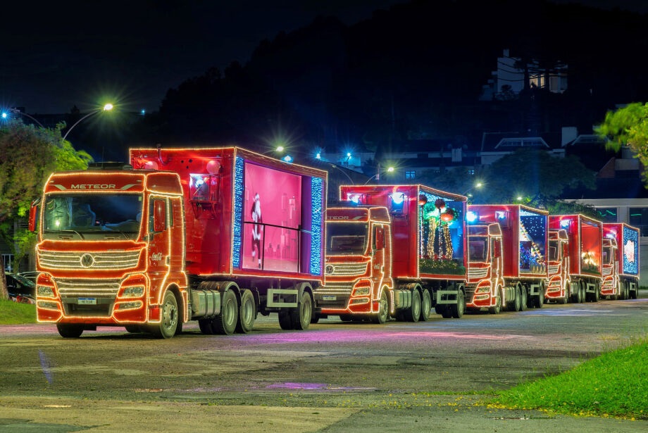 A imagem exibe uma fila de caminhões vermelhos, decorados com luzes de Natal brilhantes, estacionados em uma rua à noite. Cada caminhão possui uma tela em seu reboque, exibindo cenas temáticas de Natal, como figuras humanas e paisagens festivas. As luzes contornam os veículos, criando um espetáculo visual vibrante contra o fundo escuro da noite e as colinas ao longe. O gramado verde e as árvores adicionam um toque natural à cena, enquanto a iluminação artificial dos caminhões domina a composição.