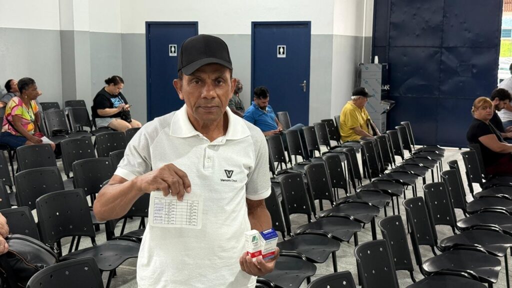 A imagem mostra um homem mais velho, vestindo uma camisa polo branca com um logo preto e um boné preto, segurando um papel com informações e duas caixas pequenas em suas mãos. Ele está em primeiro plano, olhando diretamente para a câmera. Ao fundo, há várias fileiras de cadeiras pretas vazias e algumas pessoas sentadas, que parecem estar em uma sala de espera ou local de evento. As paredes são cinzas e azuis, e há duas portas azuis com símbolos de banheiro no fundo. O ambiente sugere um local público, possivelmente uma clínica, posto de saúde ou centro comunitário.