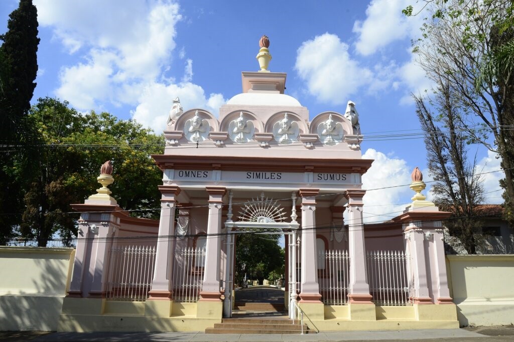 A imagem mostra um portão ornamentado, predominantemente em tons de rosa e amarelo. O portão é emoldurado por colunas e encimado por uma cúpula com um ornamento em forma de chama. Acima da entrada principal, em letras maiúsculas brancas, está inscrita a frase em latim "OMNES SIMILES SUMUS", que se traduz como "Somos todos iguais". A parte superior do portão apresenta relevos de anjos em arco, com coroas flamejantes em cada lado. A estrutura é decorada com cercas de ferro branco e uma parede baixa de cor creme. Ao fundo, através da abertura do portão, vislumbra-se um caminho que leva a uma área arborizada, possivelmente um cemitério ou um parque, com o céu azul e nuvens brancas ao fundo. A arquitetura sugere um estilo clássico com influências barrocas ou art nouveau.