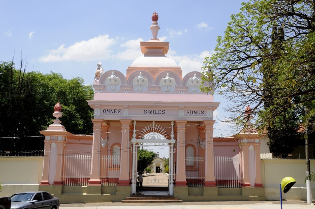 A imagem mostra a entrada de um cemitério, com um portão principal imponente e decorado em tons de rosa. O portão é encimado por uma cúpula e adornado com figuras de anjos e a inscrição "OMNES SIMILES SUMUS", que em latim significa "Todos somos iguais". Pilastras cor-de-rosa sustentam a estrutura, e grades brancas ladeiam a entrada. Árvores frondosas e um céu azul com nuvens completam a cena, criando um ambiente de serenidade. Ao lado da entrada principal, há um muro branco e uma cerca de arame farpado, sugerindo a proteção do local. Um carro estacionado na rua e um telefone público amarelo em primeiro plano adicionam um toque de realismo à imagem.