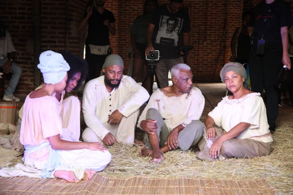 A imagem mostra um grupo de cinco pessoas sentadas no chão, que está coberto por palha e esteiras de bambu, em um ambiente que parece ser rústico, com paredes de tijolos aparentes e estrutura de madeira ao fundo. Elas vestem roupas simples e folgadas, com tons claros, que lembram trajes históricos, talvez relacionados a um período difícil, como a escravidão. Alguns usam lenços ou turbantes na cabeça. As expressões das pessoas são sérias e contemplativas, transmitindo um clima de reflexão ou tristeza. À esquerda, duas mulheres jovens estão com o olhar voltado para baixo; ao centro, um homem com barba e um lenço na cabeça também olha para baixo, enquanto um homem mais velho, ao centro-direita, olha para o lado com expressão pensativa. À direita, uma mulher com um lenço cinza na cabeça olha diretamente para a câmera com uma expressão neutra. No fundo, há outras pessoas, possivelmente parte da equipe de filmagem, uma delas segurando uma câmera. A presença desses equipamentos e pessoas sugere que a cena é parte de uma produção audiovisual, talvez um filme ou peça teatral, ambientada em um cenário histórico e dramático. A iluminação é suave, com tons terrosos predominando, reforçando o clima sóbrio da cena.