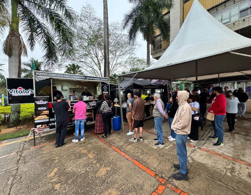 A imagem mostra uma cena externa com várias barracas de comida e pessoas. No centro, há um food truck com a marca "Vitalo's buffet & eventos", oferecendo um buffet completo para casamentos, aniversários e eventos. O food truck exibe um menu com opções como "HOT DOG" e "LANCHE DE HAMBURGUER". Várias pessoas estão em fila para comprar comida, enquanto outras conversam e circulam pelo local. Ao fundo, há um prédio com arquitetura moderna e palmeiras, sugerindo um ambiente urbano ou de evento ao ar livre. Uma grande tenda branca cobre parte da área onde as pessoas estão reunidas. A atmosfera parece ser de um evento ou feira gastronômica.