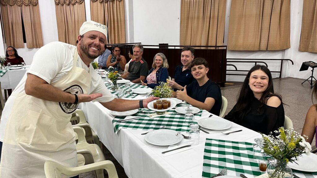 A imagem mostra um homem sorridente usando um avental branco e um chapéu de chef, apresentando uma bandeja com pequenos copos de uma sobremesa marrom para uma mesa de jantar. A mesa está posta com toalhas xadrez verdes e brancas, pratos brancos, talheres e copos de água. Há um arranjo de flores amarelas e brancas no centro da mesa. Várias pessoas estão sentadas à mesa, aproveitando a refeição. Ao fundo, cortinas marrons cobrem as janelas e uma estrutura de metal com um suporte para microfone pode ser vista. A atmosfera geral é festiva e acolhedora, sugerindo um evento de jantar ou celebração.