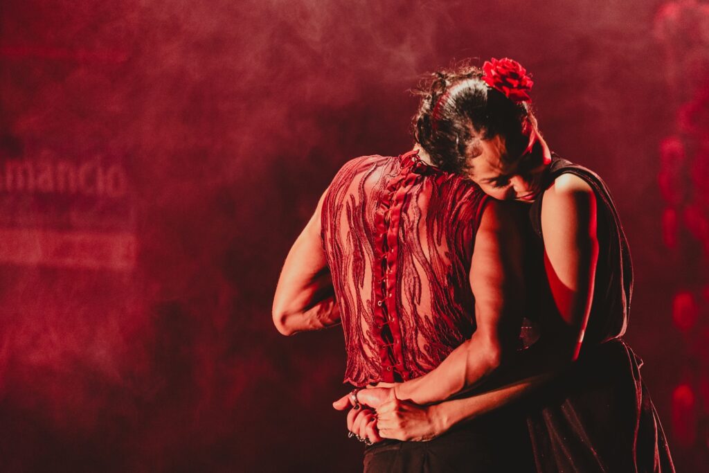 A imagem mostra dois bailarinos em um abraço íntimo, com iluminação dramática em tons de vermelho. Um dos bailarinos, com uma rosa vermelha no cabelo, apoia a cabeça no ombro do outro, que veste uma blusa translúcida com detalhes em renda e amarração nas costas. A atmosfera é carregada de emoção e movimento, sugerindo uma performance de dança intensa. Ao fundo, um texto em letras claras, possivelmente parte do cenário, adiciona um elemento visual à cena.