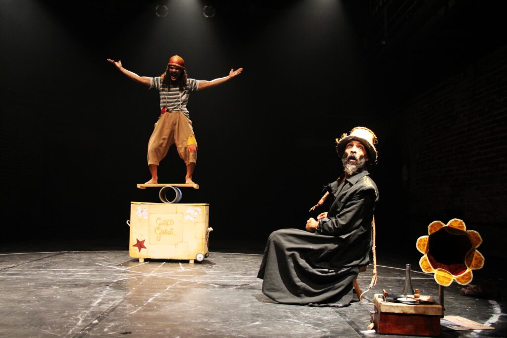 A imagem mostra uma cena de teatro ou circo em um palco escuro, com iluminação focada nos dois atores. À esquerda, um artista está em pé sobre uma prancha de madeira equilibrada em um cilindro, que por sua vez está apoiado sobre uma caixa com rodinhas decorada com uma estrela vermelha e desenhos infantis. Ele usa uma camiseta listrada, calça larga bege com um remendo amarelo e um gorro vermelho, e está com os braços abertos, sorrindo de forma expressiva, transmitindo energia e alegria. À direita, um segundo artista está sentado em um banco, vestindo um casaco escuro e um chapéu desgastado. Sua expressão é de surpresa ou admiração, olhando para o artista que está equilibrando-se. Perto dele, há alguns objetos, incluindo um toca-discos antigo com um disco de vinil, uma garrafa de vidro e um objeto que parece uma luminária ou um adereço decorativo. O palco é simples, com um fundo escuro e detalhes como uma parede de tijolos à direita, que adiciona profundidade à cena. O ambiente sugere um espetáculo intimista de circo ou teatro de rua, focado na interação e no equilíbrio entre os personagens. O texto “Circo Goast” aparece na caixa, indicando possivelmente o nome do espetáculo ou do grupo.