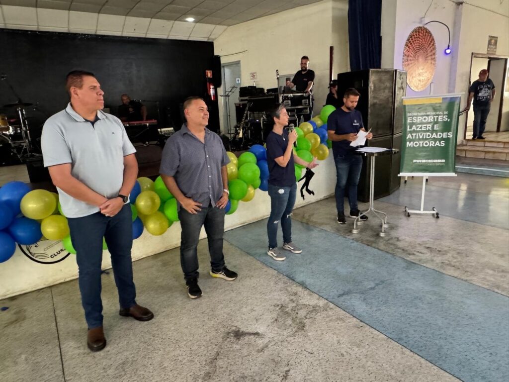 A imagem mostra um evento com várias pessoas em frente a um palco. Há balões azuis e amarelos decorando o local. No palco, há equipamentos de som e músicos. À frente do palco, quatro pessoas estão em destaque. Duas delas, homens, estão em pé, de frente para a plateia. Uma mulher, segurando um microfone, fala para o público, enquanto um homem ao seu lado lê um documento. Ao fundo, um homem está em pé, próximo a uma porta, vestindo uma camiseta com a inscrição "JOBS". Há também um banner com a inscrição "SECRETARIA MUNICIPAL DE ESPORTES, LAZER E ATIVIDADES MOTORAS".