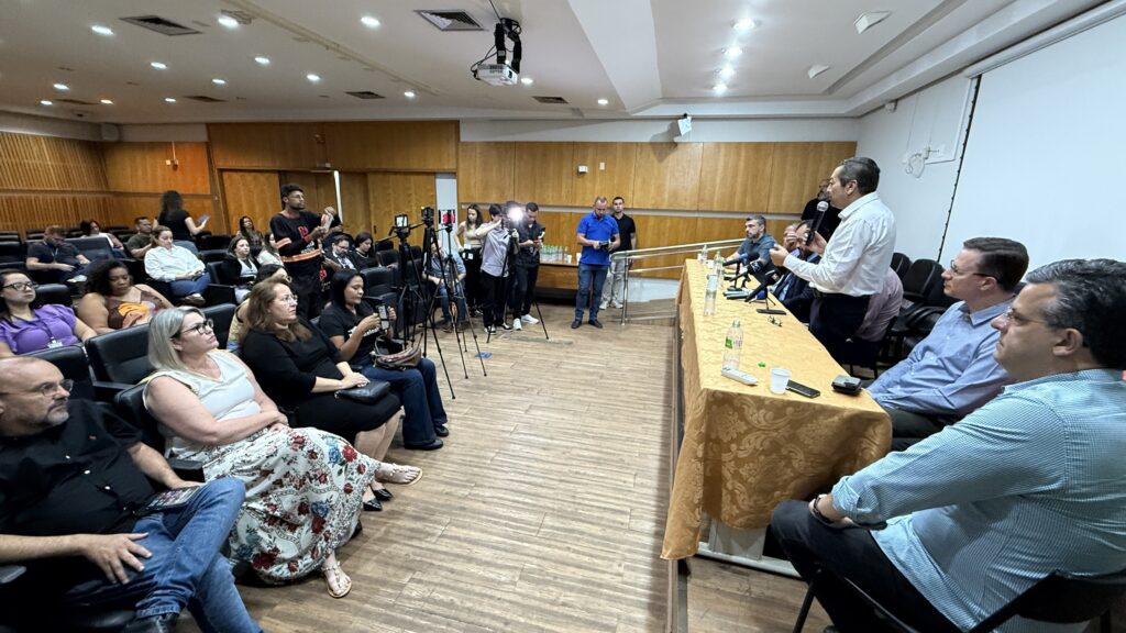 A imagem retrata uma sala de conferências com um público sentado em cadeiras e um grupo de pessoas em pé perto de uma mesa no fundo, onde um palestrante está falando ao microfone. Vários fotógrafos e cinegrafistas com equipamentos de gravação estão posicionados perto da mesa, indicando um evento de mídia ou uma conferência de imprensa. O público está composto por homens e mulheres de diversas idades, a maioria deles olhando para o palestrante ou para o lado. Alguns estão segurando seus celulares, possivelmente para tirar fotos ou gravar o evento. As cadeiras são escuras e dispostas em fileiras, e o ambiente parece formal, com painéis de madeira nas paredes e iluminação de teto. No centro da imagem, um homem de camisa branca está de pé, segurando um microfone e falando para a plateia. Ele está posicionado atrás da mesa principal, onde estão sentados outros homens, possivelmente convidados ou participantes do evento. A mesa está coberta com uma toalha dourada com um padrão texturizado e há garrafas de água e outros objetos sobre ela. No fundo, um grupo de pessoas, incluindo os cinegrafistas e fotógrafos, está em pé. Alguns deles parecem estar interagindo com o palestrante ou com os convidados na mesa. Há um projetor pendurado no teto, sugerindo que o evento pode envolver apresentações visuais. A atmosfera geral da imagem sugere um evento de importância, possivelmente com cobertura da mídia.