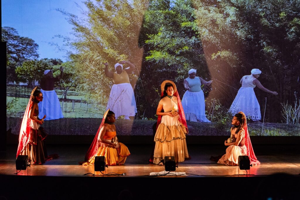 A imagem mostra uma performance teatral em um palco iluminado. Ao fundo, uma projeção exibe uma paisagem rural com árvores e figuras de mulheres em movimento, vestidas com roupas tradicionais, incluindo lenços na cabeça e saias longas. Em primeiro plano, cinco artistas estão presentes. No centro, uma artista com um chapéu de palha e um véu vermelho, segura um objeto dourado e veste um longo vestido de época em tons de creme e bege. À sua esquerda, outra artista, com um véu semelhante e um traje laranja e vermelho, ajoelha-se segurando um vaso. À direita, uma terceira artista, com o cabelo preso em tranças e um véu vermelho, também ajoelhada, segura um objeto semelhante. Nas extremidades do palco, outras duas artistas estão em pé, vestidas com trajes que lembram as figuras projetadas ao fundo, com véus e saias longas em tons de vermelho e marrom. A iluminação do palco cria um ambiente dramático, destacando os artistas em primeiro plano e a projeção ao fundo.